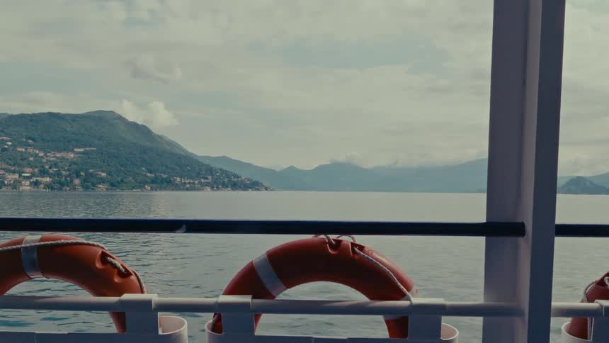 A serene view from a ferry crossing a large mountain lake on a cloudy day, with orange lifebuoys in the foreground