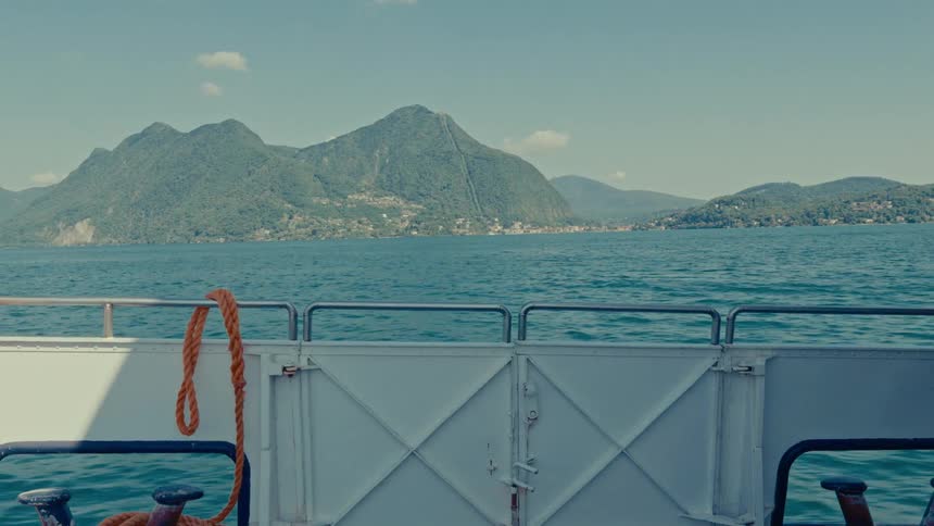 The view from the back of a ferry as it peacefully crosses a stunning blue lake surrounded by lush green mountains on a sunny day
