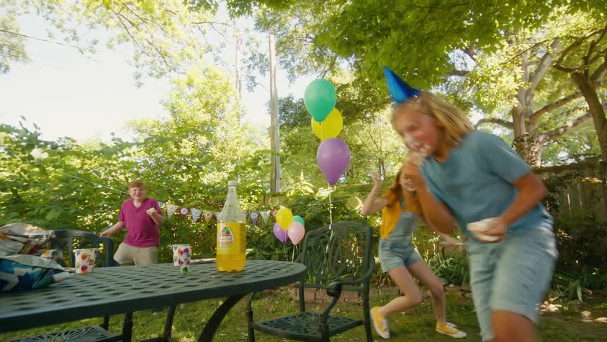 Three happy children with cake on their faces playfully chase each other during a sunny backyard birthday party