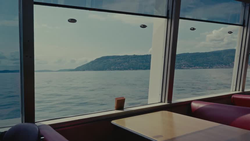 A scenic view from inside a moving ferry shows the calm water and distant tree-covered hills passing by the window