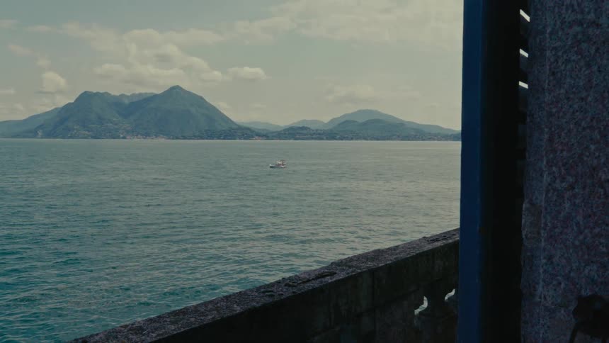 A serene view from a stone balcony captures a lone boat sailing on a vast lake with lush green mountains in the background
