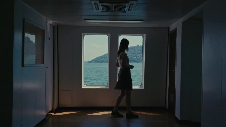 A contemplative woman in silhouette gazes out the window of a moving ferry, watching the sunlit water and hills pass by