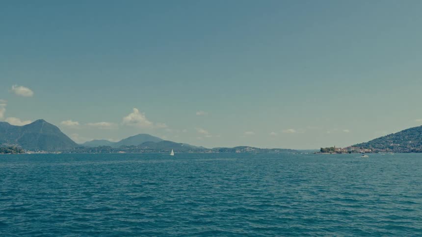 A tranquil wide shot of a vast blue lake, with a lone sailboat gliding across the water against a backdrop of majestic mountains and a clear sky