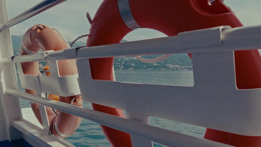 A bright orange life preserver hangs from the white railing of a moving boat, with a scenic view of the sea and mountains in the background