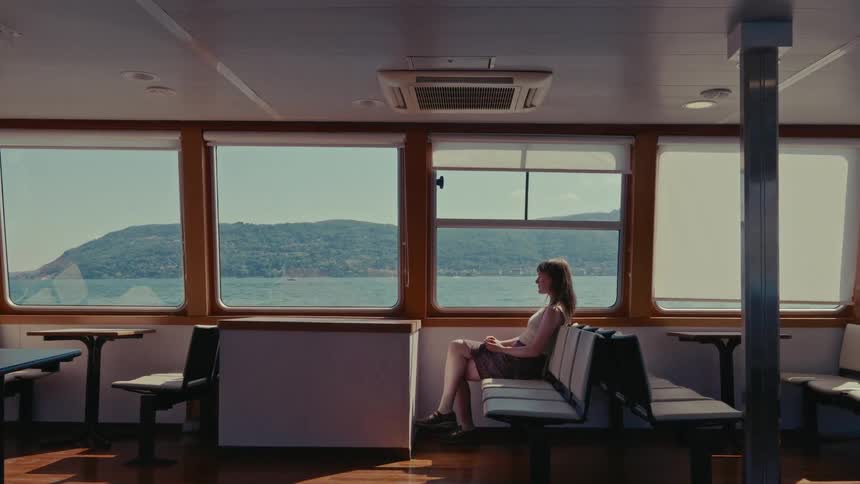 A woman sits alone inside a moving ferry, looking out the window at the serene water and lush green coastline