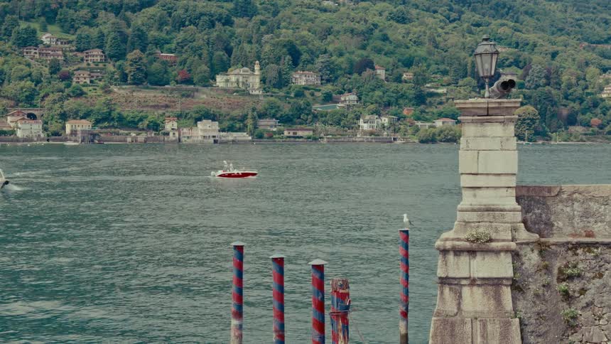Boats cruise on a scenic European lake, passing by a stone pier and colorful striped mooring poles with a verdant hillside town in the background