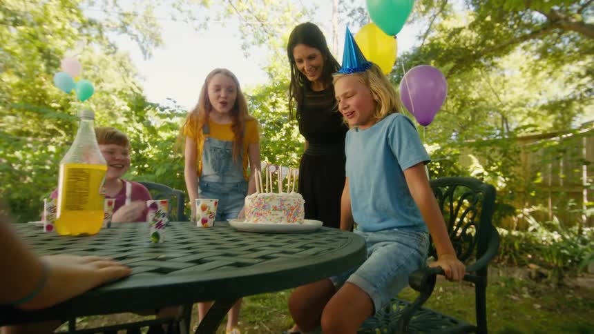 A happy boy makes a wish and blows out his birthday candles during a backyard party with his friends and mom