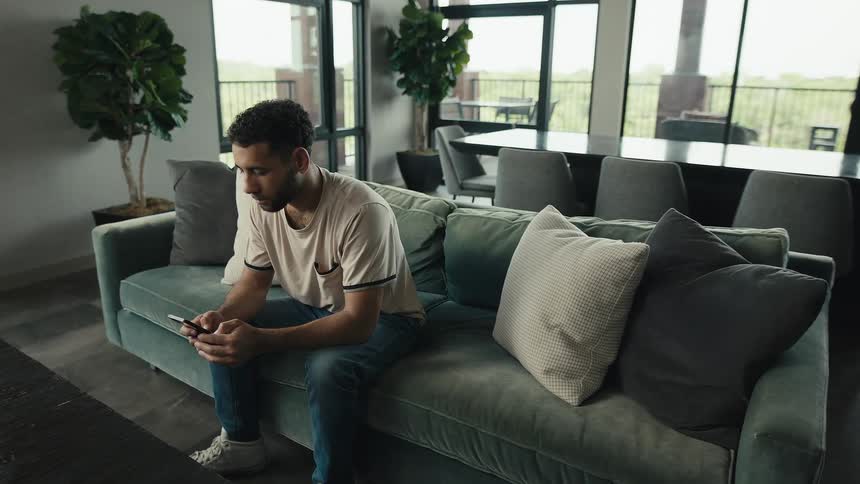 A man sits alone on a stylish couch in a modern living area, focused on scrolling through his smartphone