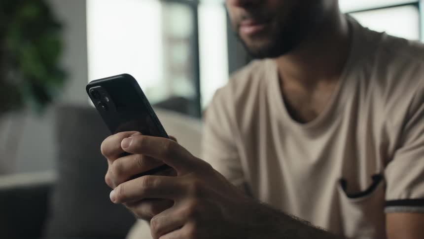 A close-up slow-motion shot of a young man smiling as he scrolls on his smartphone while relaxing at home