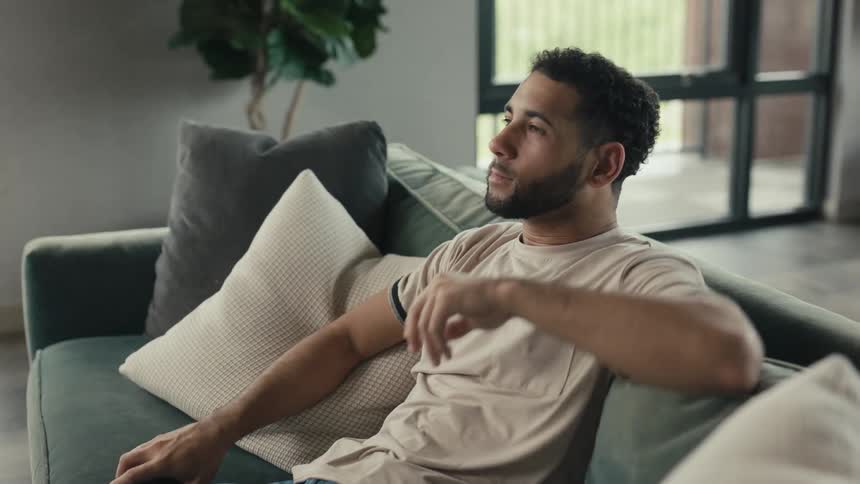 A relaxed young man scrolls on his smartphone before putting it down to unwind on the couch in his living room