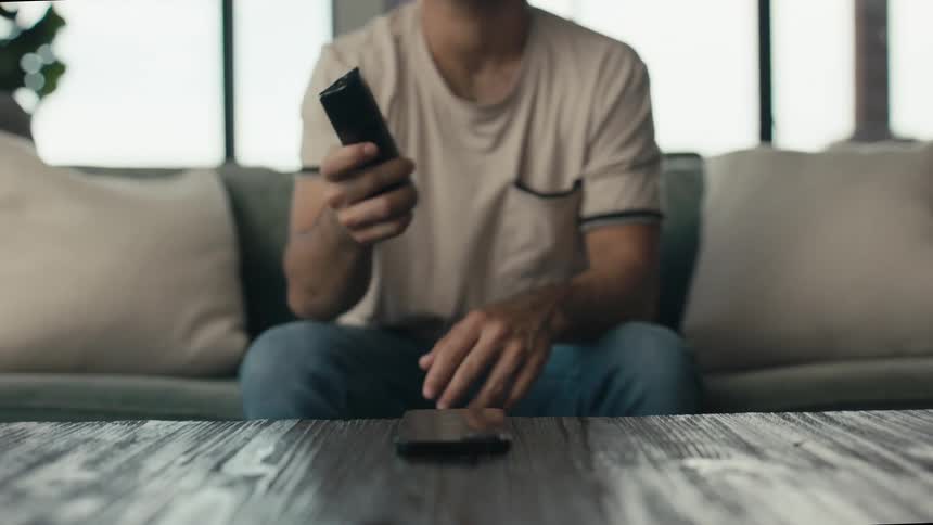 A man puts down his phone and picks up the TV remote in slow motion to relax and watch TV