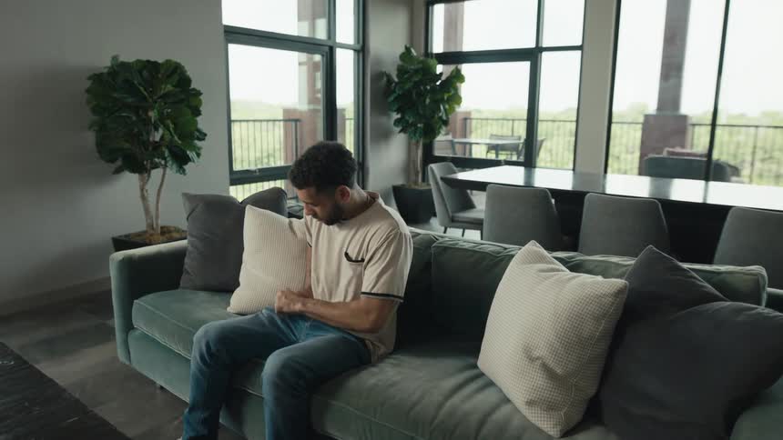A man walks into a modern living room, sits down on the sofa, and scrolls on his smartphone