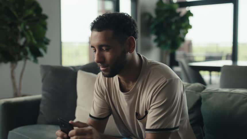 A young man casually pulls out his smartphone to browse while relaxing on the couch in his modern, sunlit living room