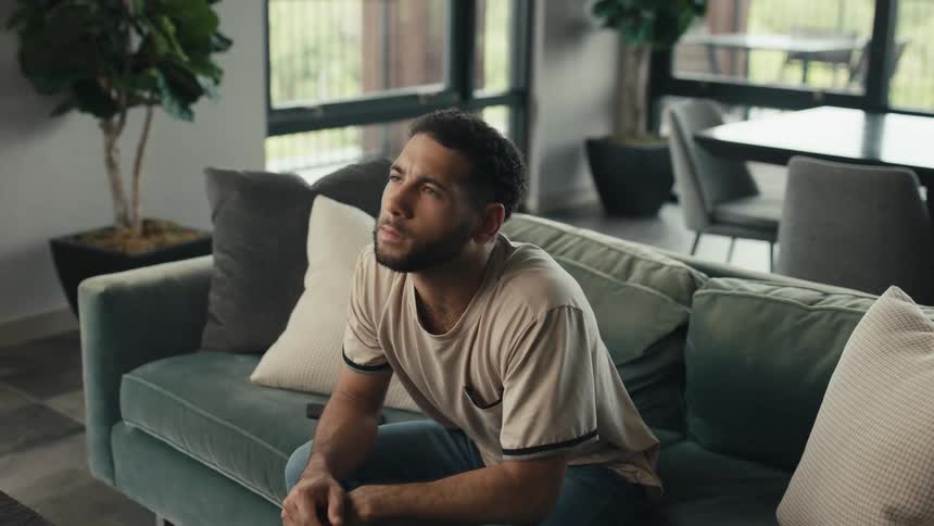 Young man puts down his phone while sitting on a couch and reflects quietly in his modern living room