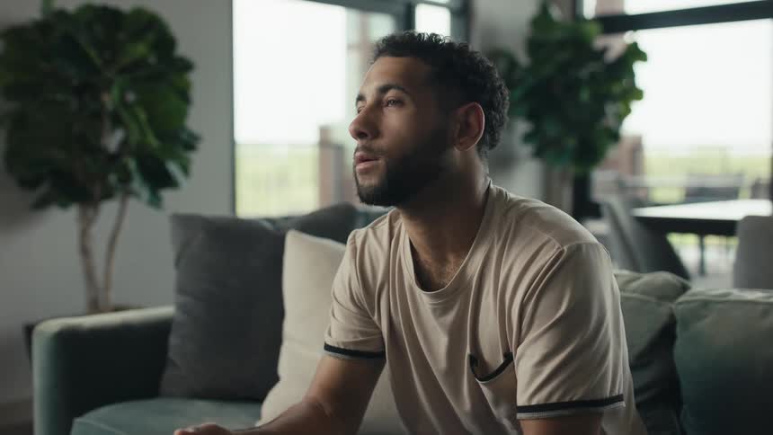 A relaxed young man puts down his smartphone and uses the remote to watch television while sitting on the couch in his modern living room