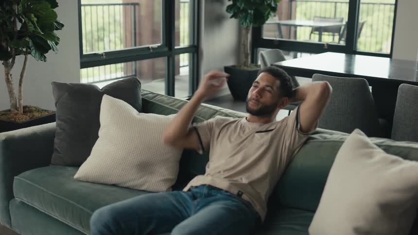 Happy young man puts his phone down and leans back to relax on the couch in his modern apartment
