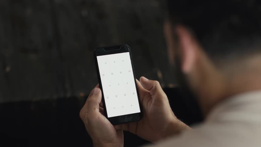 Over the shoulder shot of a man's hands holding a smartphone with a blank white screen and tracking markers for compositing