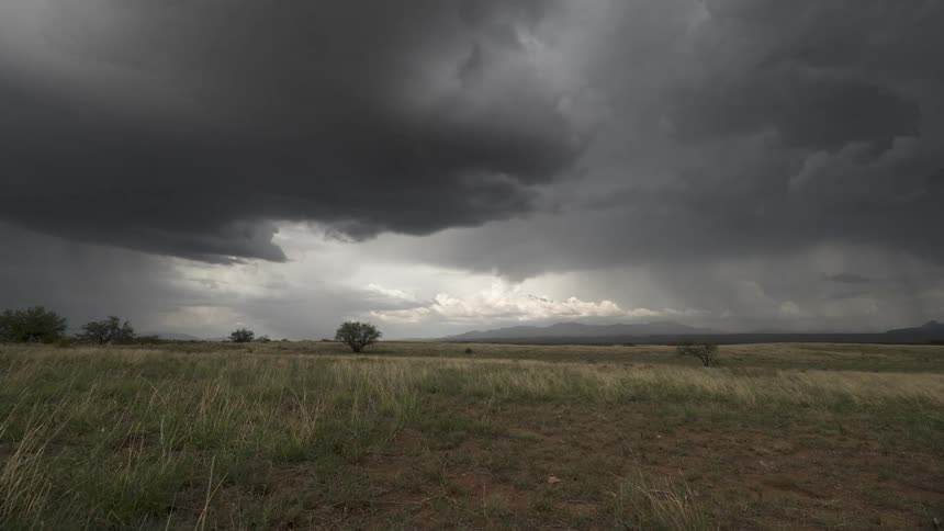 A dramatic time-lapse captures ominous dark storm clouds gathering and releasing rain over a vast grassy plain