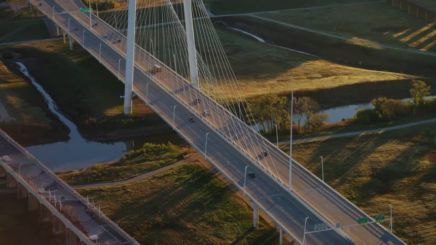 An aerial view shows traffic driving across a modern cable-stayed bridge over a river during a beautiful golden sunrise