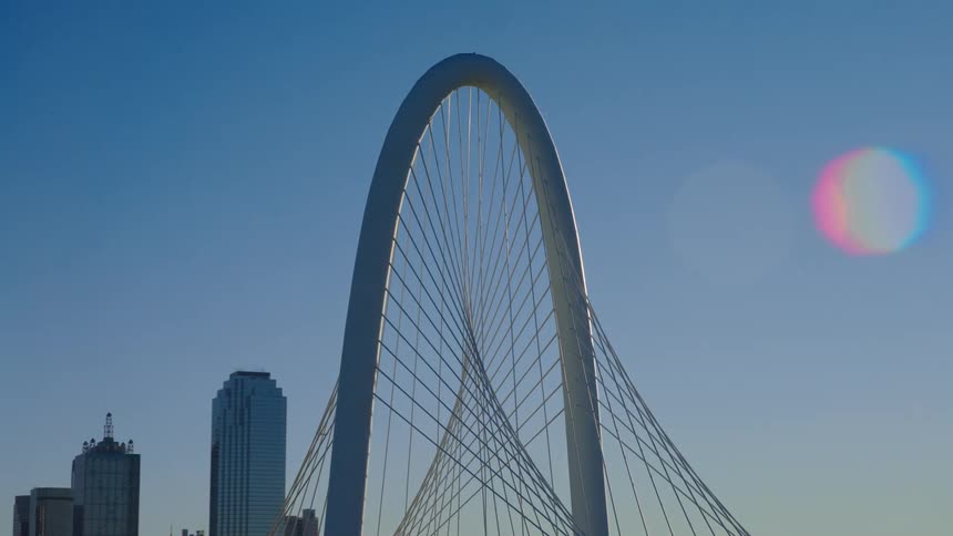 Slow zoom out from the iconic Margaret Hunt Hill Bridge arch against a sunlit blue sky, revealing the Dallas, Texas skyline