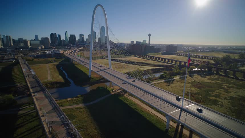 A stunning aerial pull-back shot reveals the Margaret Hunt Hill Bridge and the Dallas skyline under a bright morning sun