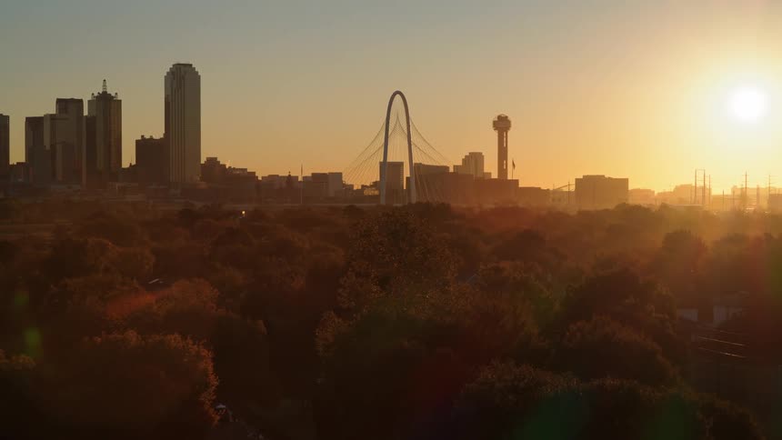 Slow aerial pull-back shot of the Dallas, Texas skyline with iconic landmarks during a hazy golden hour sunset