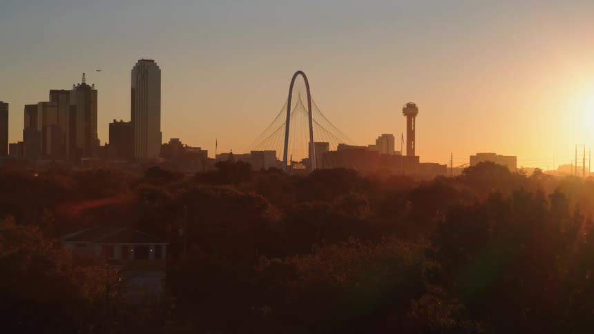The iconic Dallas skyline is silhouetted against a beautiful, warm sunrise in a slow and steady aerial pull-back shot over a treelined neighborhood