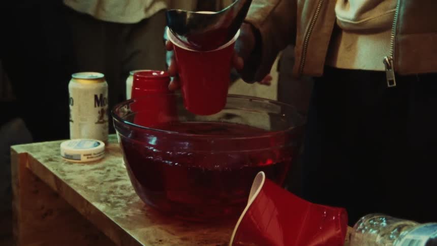 A close-up of someone ladling red punch from a large glass bowl into a plastic red cup at a house party