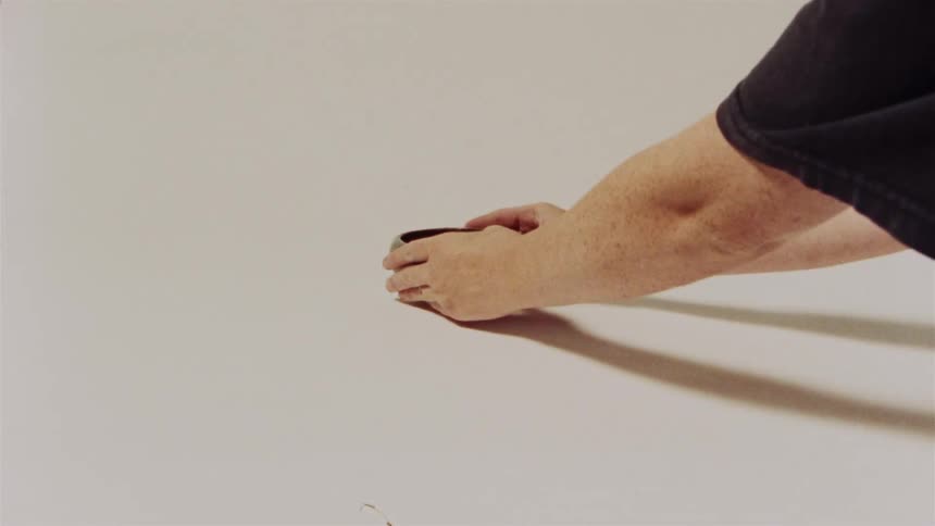 A person's hands carefully place a small, dark ceramic bowl onto a plain white surface