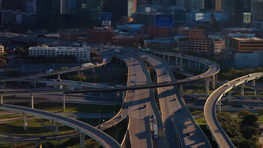 Aerial view of a sprawling highway interchange with steady traffic flowing in front of the Dallas, Texas skyline at golden hour