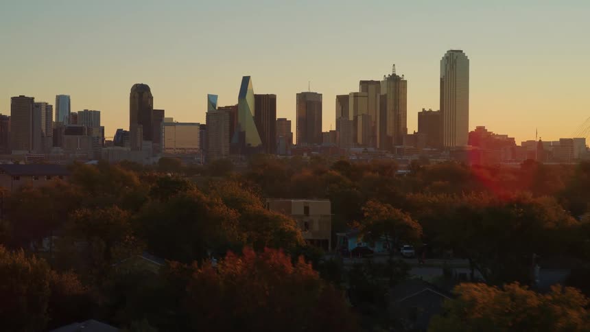 A cinematic aerial drone shot rises over a quiet suburban neighborhood to reveal the Dallas, Texas skyline at sunrise