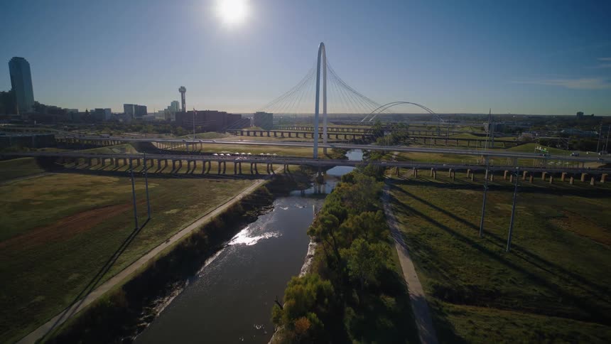 A soaring aerial shot reveals the Dallas, Texas skyline, featuring the iconic Margaret Hunt Hill Bridge over the Trinity River on a bright, sunny morning