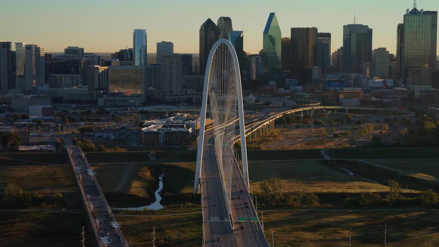 The iconic Margaret Hunt Hill Bridge stands before the downtown Dallas skyline, illuminated by the warm light of sunrise in this sweeping aerial shot