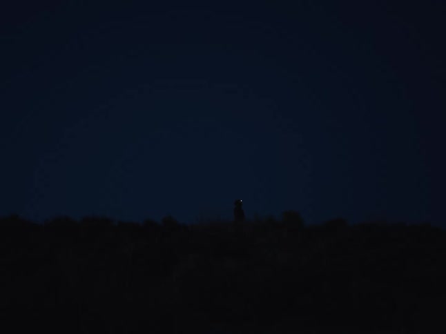 A silhouette of a person standing on a hill at night turns on their headlamp