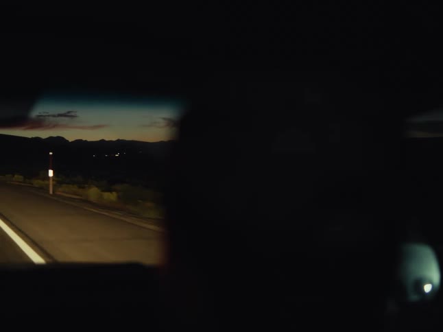 A driver's silhouette is seen against the reflection of a desert highway at dusk in the rearview mirror