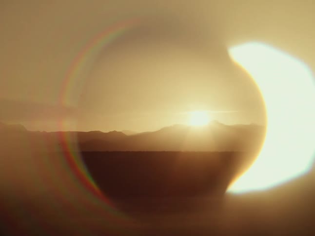 An artistic and abstract view of a golden sunset over a silhouetted mountain range, framed by a surreal, circular lens flare