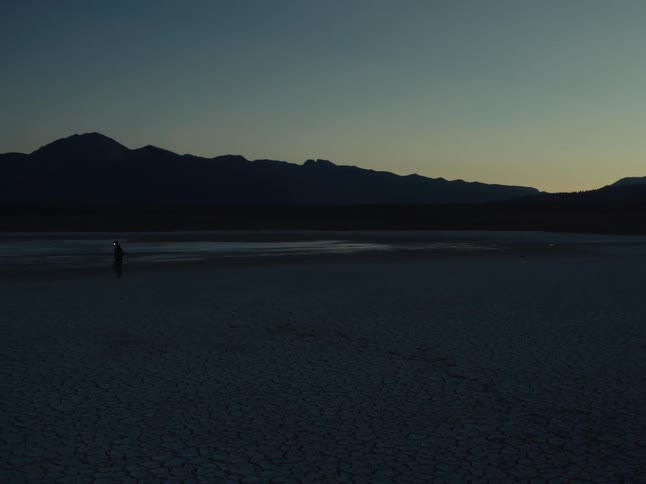 A solitary traveler wearing a headlamp walks across a vast, dark, and cracked desert landscape at twilight