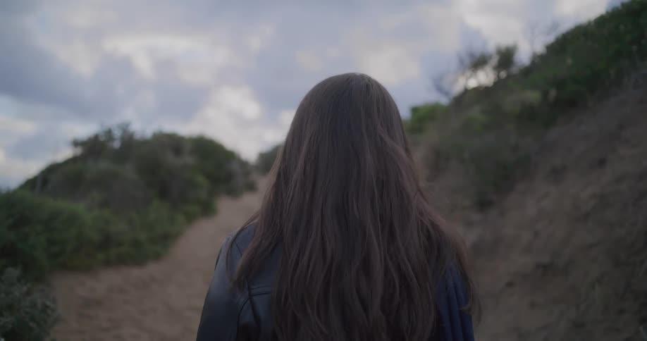A slow-motion, over-the-shoulder shot of a young woman with long brown hair standing on a sandy path leading up a hill on a cloudy day