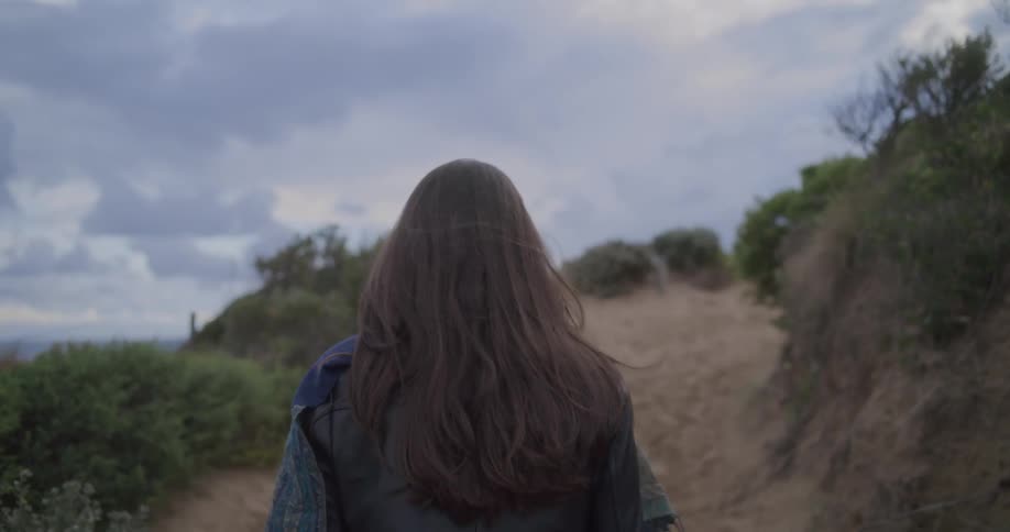 A woman with long dark hair walks along a sandy path on a windy and overcast day, seen from behind in slow motion