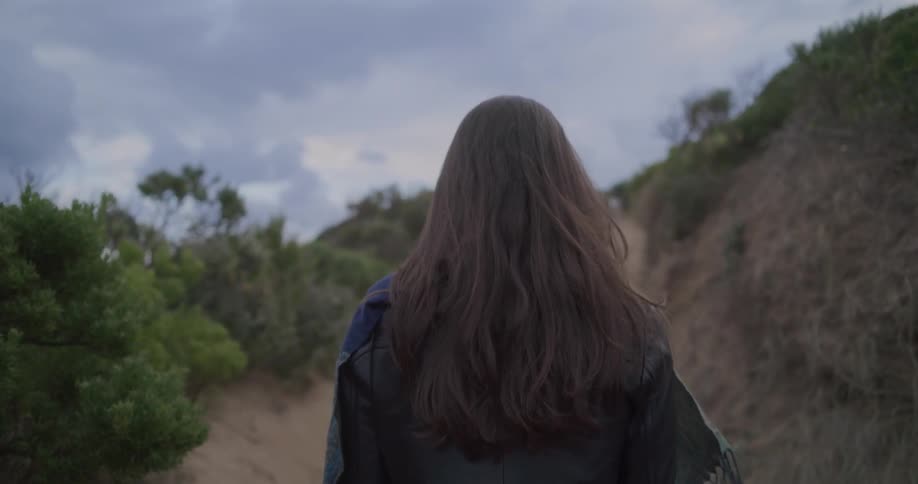 A slow-motion shot follows a woman with long brown hair as she walks up a sandy path on a cloudy day