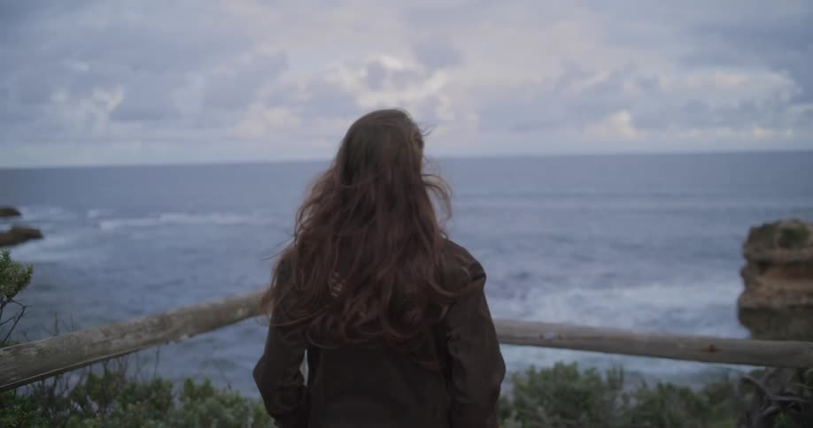 Slow motion shot from behind of a person with long hair blowing in the wind as they look out over the ocean from a cliff on a cloudy day