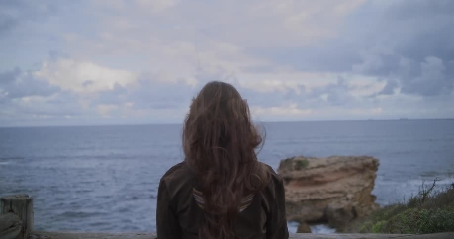 Slow motion shot of a woman with long hair blowing in the wind as she looks out over the ocean on an overcast day