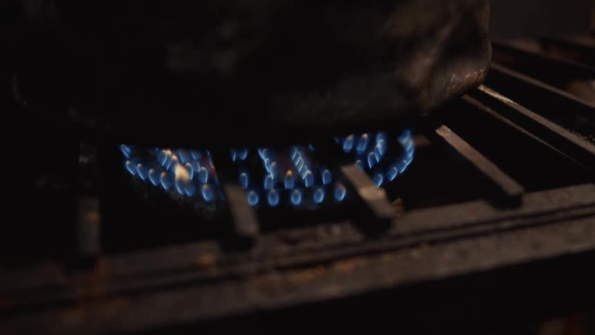 A close-up of blue gas flames from a stovetop burner heating a dark pot in a dimly lit setting