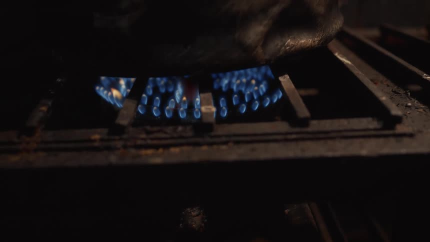A close-up shot of the blue flames of a gas stove burner heating a pot in a dark kitchen