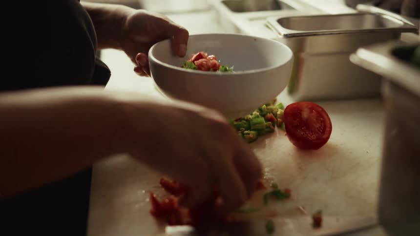 A chef in slow motion scoops freshly chopped tomatoes and peppers from a cutting board into a white bowl in a kitchen