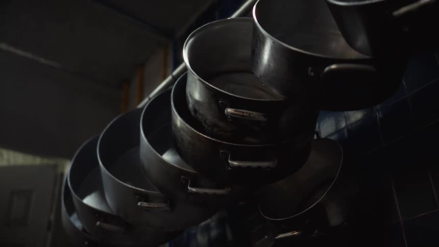 A row of large, old metal cooking pots hangs on a rack in a dark, industrial kitchen