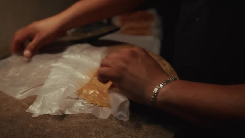 A cook pounds a piece of meat flat with a cleaver on a wooden block and wraps it in plastic in slow motion