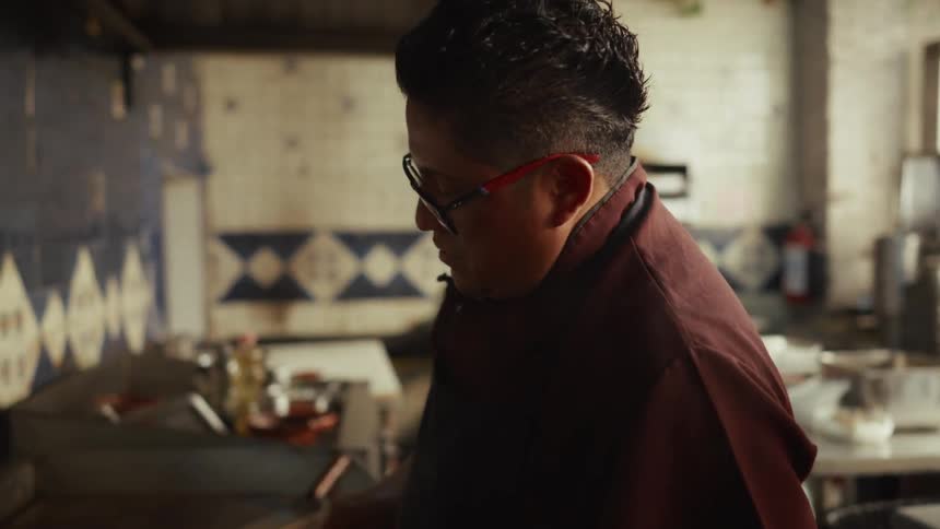A chef wearing an apron and glasses carefully prepares a meal on a large steel griddle in a commercial kitchen