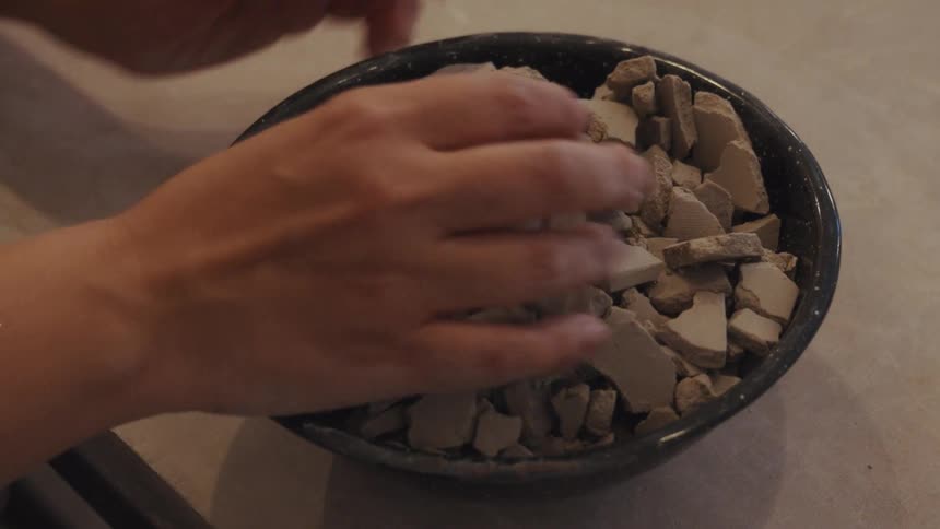 Slow motion shot of hands touching and pressing down on shards of dry clay in a black bowl