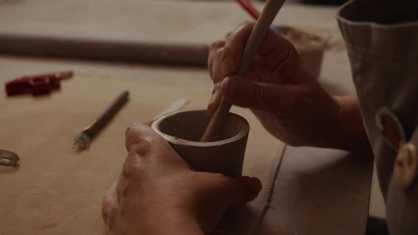 A ceramic artist carefully uses a wooden tool to smooth the inside walls of a handmade clay pot on their workbench